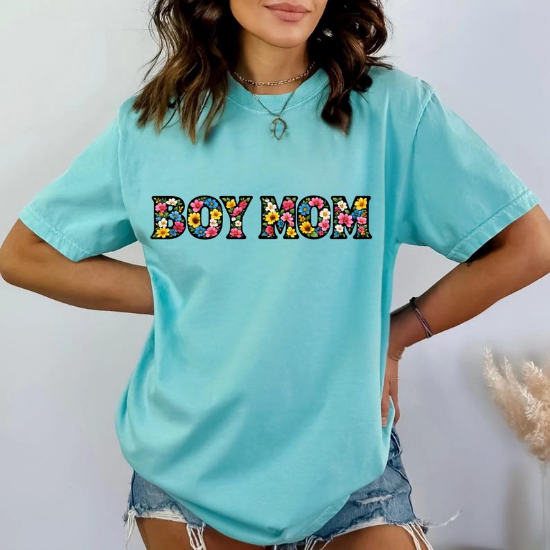 May include: A light blue t-shirt with the text "BOY MOM" in a floral design. The shirt has short sleeves and a relaxed fit. The text is in a bold, block letter style with a colorful flower pattern.