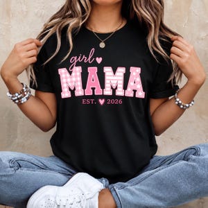 Könnte beinhalten: Ein schwarzes Kurzarm-T-Shirt mit dem rosa Schriftzug girl MAMA und einem kleinen Herzsymbol. Die Buchstaben in MAMA haben ein rosa-weißes Vichy-Karomuster und Sternakzente. Unter dem Text steht der Schriftzug EST. 2026 mit einem kleinen rosa Herz.