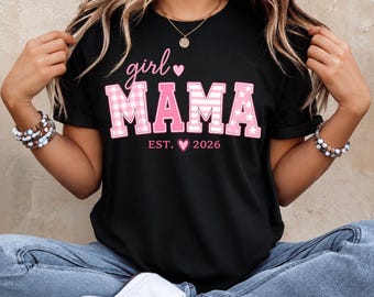 May include: A black short-sleeved t-shirt featuring the pink text girl MAMA with a small heart icon. The letters in MAMA have a pink and white gingham pattern and star accents. Below the text is the phrase EST. 2026 with a small pink heart.