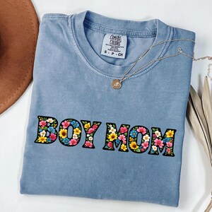 May include: A light blue Comfort Colors t-shirt with the words "BOY MOM" in a floral design. A gold necklace with a pendant is draped on the shirt. The shirt is a size small.