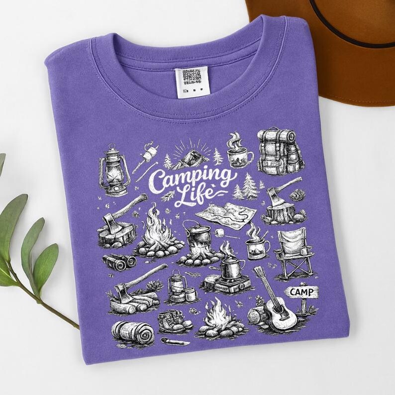 May include: A purple t-shirt featuring a camping-themed graphic. The design includes illustrations of a lantern, campfire, guitar, and the words "Camping Life." The word "CAMP" is also visible.