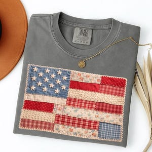 May include: Gray t-shirt with a patchwork American flag design. The flag features blue denim with white stars, and red and floral patterned fabric stripes. A gold necklace with a pendant rests on the shirt.