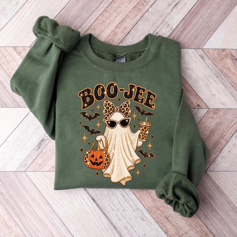 Pu&ograve; includere: Una felpa girocollo verde oliva con la grafica di un fantasma che indossa un fiocco e occhiali da sole leopardati. Il fantasma tiene in mano una zucca e un bicchiere, circondato da pipistrelli e stelle. Il testo BOO-JEE &egrave; stampato sopra.