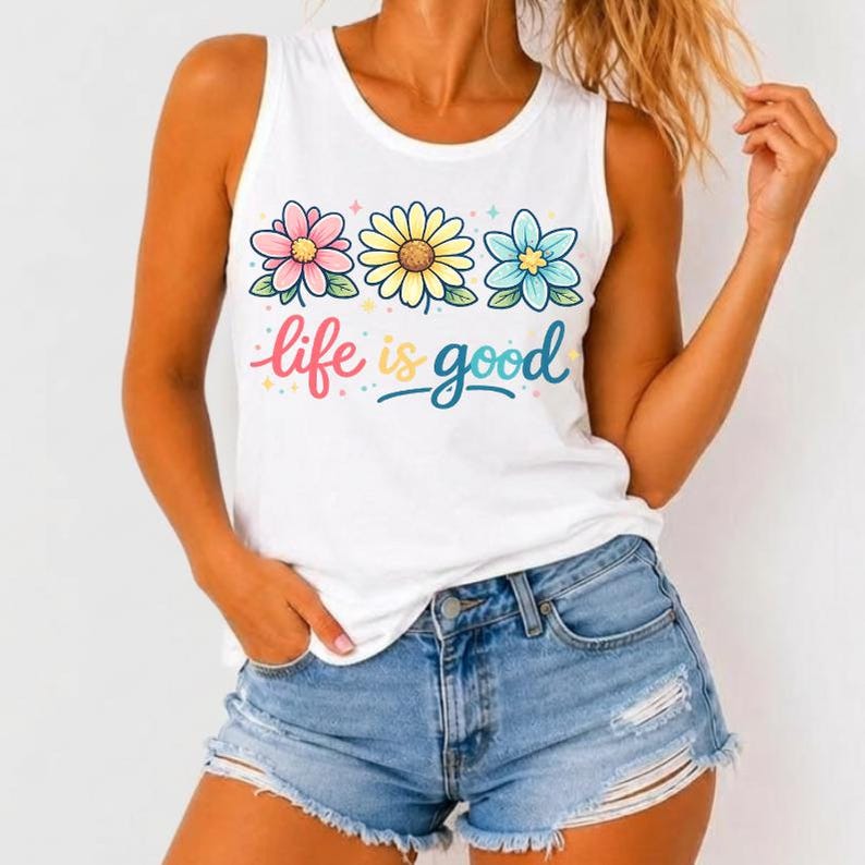 May include: White tank top with a floral design and the phrase "life is good" in colorful script. The top features three stylized flowers in pink, yellow, and blue. The model is wearing denim shorts.
