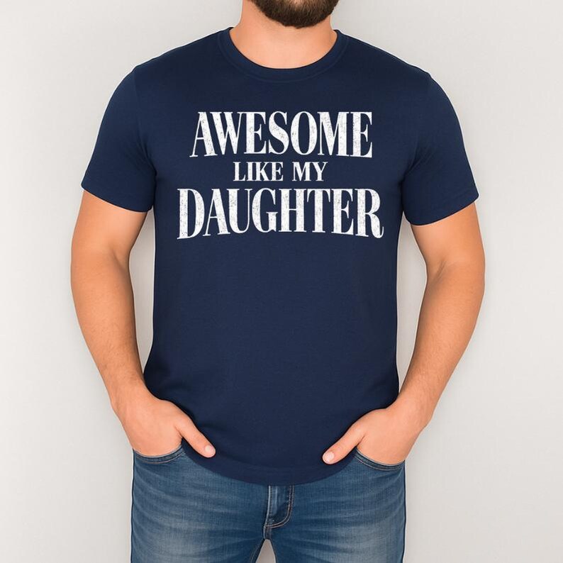 May include: A navy blue t-shirt with the white text "AWESOME LIKE MY DAUGHTER". The shirt has a crew neck and short sleeves. The person is wearing blue jeans. The text is in uppercase.