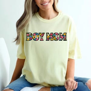 May include: A light yellow t-shirt with the text "BOY MOM" in a floral design. The letters are black with a colorful flower pattern. The shirt has short sleeves and a relaxed fit.