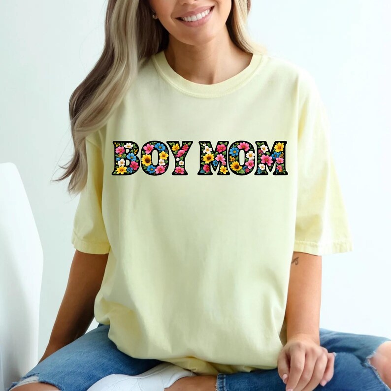 May include: A light yellow t-shirt with the text "BOY MOM" in a floral design. The letters are black with a colorful flower pattern. The shirt has short sleeves and a relaxed fit.