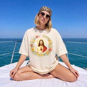 K&ouml;nnte beinhalten: Cremefarbenes T-Shirt mit einem religi&ouml;sen Bild von Jesus Christus in einem Blumenkranz. Die Grafik enth&auml;lt einen Heiligenschein und ein Herz. Das Shirt wird von einer Person auf einem Boot getragen, mit einem Leoparden-Schal und Sonnenbrille.