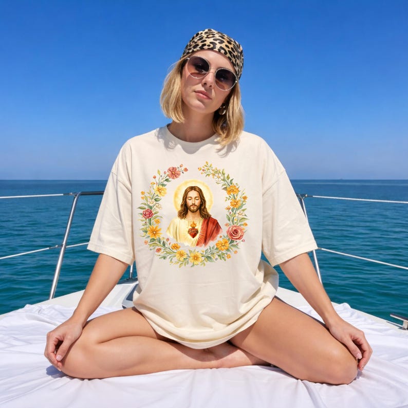 K&ouml;nnte beinhalten: Cremefarbenes T-Shirt mit einem religi&ouml;sen Bild von Jesus Christus in einem Blumenkranz. Die Grafik enth&auml;lt einen Heiligenschein und ein Herz. Das Shirt wird von einer Person auf einem Boot getragen, mit einem Leoparden-Schal und Sonnenbrille.