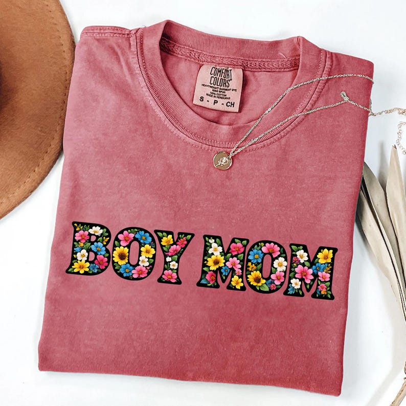 May include: A dusty rose-colored t-shirt with the words "BOY MOM" in large, black letters filled with colorful flowers. The shirt has a crew neck and short sleeves. A silver necklace with a small pendant is draped across the shirt.