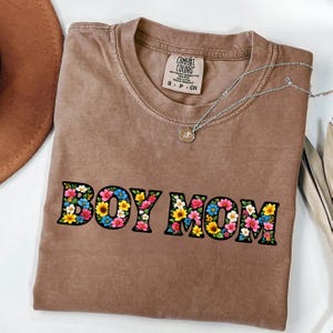 May include: A brown Comfort Colors t-shirt with the words "BOY MOM" in a floral design. The shirt has a crew neck and short sleeves. A silver necklace with a small pendant is on the shirt.