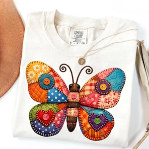 May include: White t-shirt featuring a colorful patchwork butterfly design. The butterfly has wings with various patterns, including polka dots, floral prints, and plaid, and is embellished with buttons. A gold necklace and a hat are also visible.