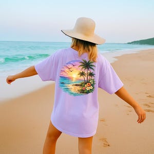 May include: A lilac t-shirt with a tropical sunset design on the back. The design features palm trees, flowers, and birds against a colourful sunset over the ocean. The person is wearing a straw hat.