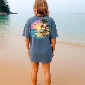 May include: A blue t-shirt with a tropical sunset design on the back. The graphic shows palm trees, flowers, and birds against a sunset sky over the ocean. The shirt is a solid blue colour.