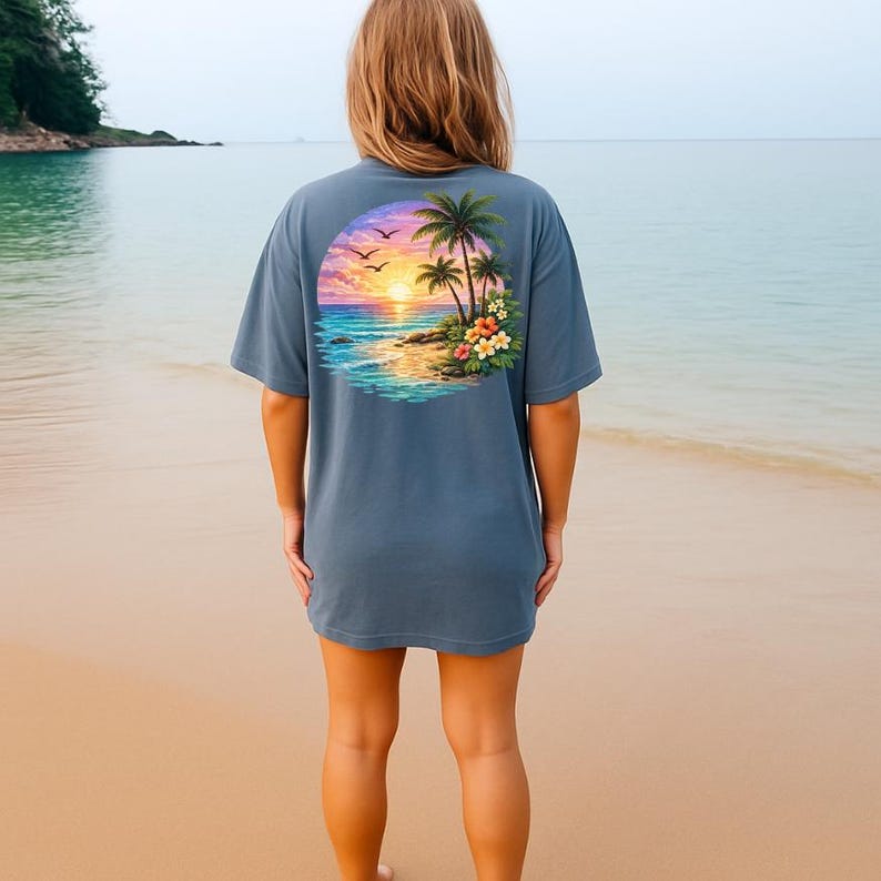 May include: A blue t-shirt with a tropical sunset design on the back. The graphic shows palm trees, flowers, and birds against a sunset sky over the ocean. The shirt is a solid blue colour.