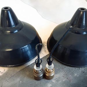 May include: Two dark gray metal pendant light shades with matching bulb sockets. The shades have a glossy finish and a classic, industrial design. The bulb sockets are a mix of metal tones.