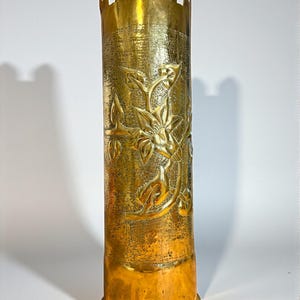 Op de afbeelding: Een hoge, cilindrische messing vaas met een bloemmotief. De vaas heeft een kroonvormige bovenkant met boogvormige uitsparingen. Het messing heeft een gepolijste, gouden afwerking.