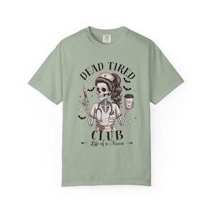 May include: A sage green t-shirt featuring a graphic of a skeleton nurse wearing a cap and stethoscope. The skeleton holds a syringe and a coffee cup. Text reads DEAD TIRED CLUB and Life of a Nurse, surrounded by small bats and star icons.