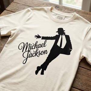 Könnte beinhalten: Cremefarbenes T-Shirt mit einer schwarzen Silhouette von Michael Jackson in einer Tanzpose. Die Worte "Michael Jackson" sind in einer Schreibschrift geschrieben. Das Shirt hat einen Rundhalsausschnitt und ein kleines Etikett mit der Aufschrift "King of Pop".