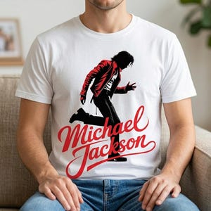 Könnte beinhalten: Weißes T-Shirt mit einer rot-schwarzen Grafik von Michael Jackson in Laufpose. Der Text "Michael Jackson" ist in roter Schrift darunter geschrieben. Das Shirt hat einen klassischen Rundhalsausschnitt.