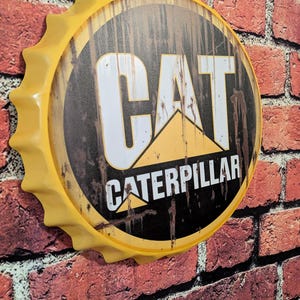 May include: A yellow metal sign in the shape of a bottle cap, featuring the CAT Caterpillar logo. The sign is mounted on a red brick wall. The logo includes the word "CAT" in white letters and "CATERPILLAR" in black.