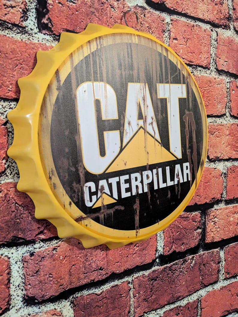 May include: A yellow metal sign in the shape of a bottle cap, featuring the CAT Caterpillar logo. The sign is mounted on a red brick wall. The logo includes the word "CAT" in white letters and "CATERPILLAR" in black.