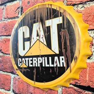 May include: A large, yellow bottle cap-shaped sign with the CAT Caterpillar logo. The sign has a black background with the logo in white and yellow. The sign is mounted on a red brick wall.