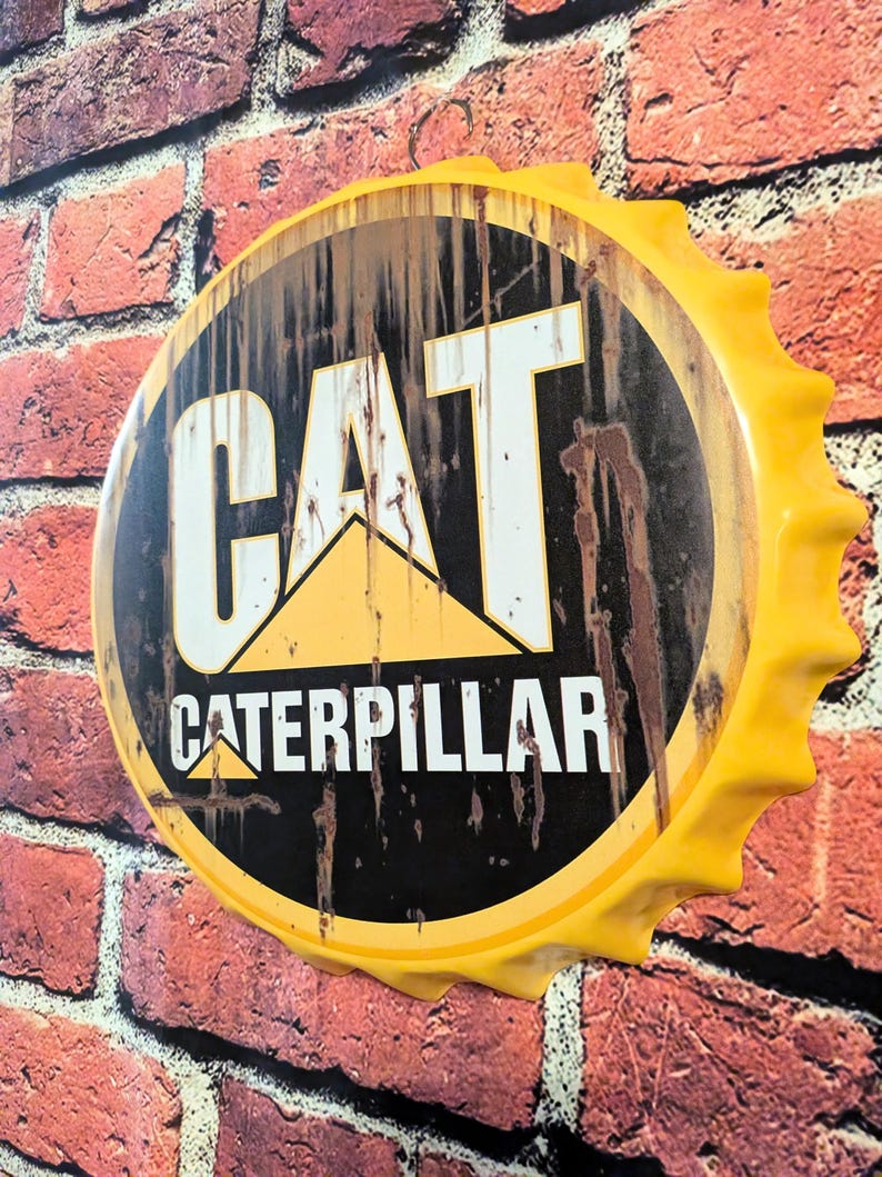 May include: A large, yellow bottle cap-shaped sign with the CAT Caterpillar logo. The sign has a black background with the logo in white and yellow. The sign is mounted on a red brick wall.