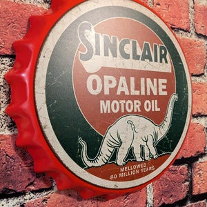May include: A red, bottle cap-shaped sign with the Sinclair Opaline Motor Oil logo. The sign features a dinosaur illustration and text in white and green on a red and white background. The sign is mounted on a brick wall.