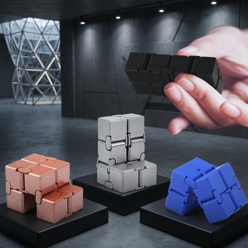 May include: An assortment of fidget toys in various colors, including copper, silver, and blue, displayed on black pedestals. A black fidget toy is held in a hand. The toys are made of connected cubes that can be manipulated.