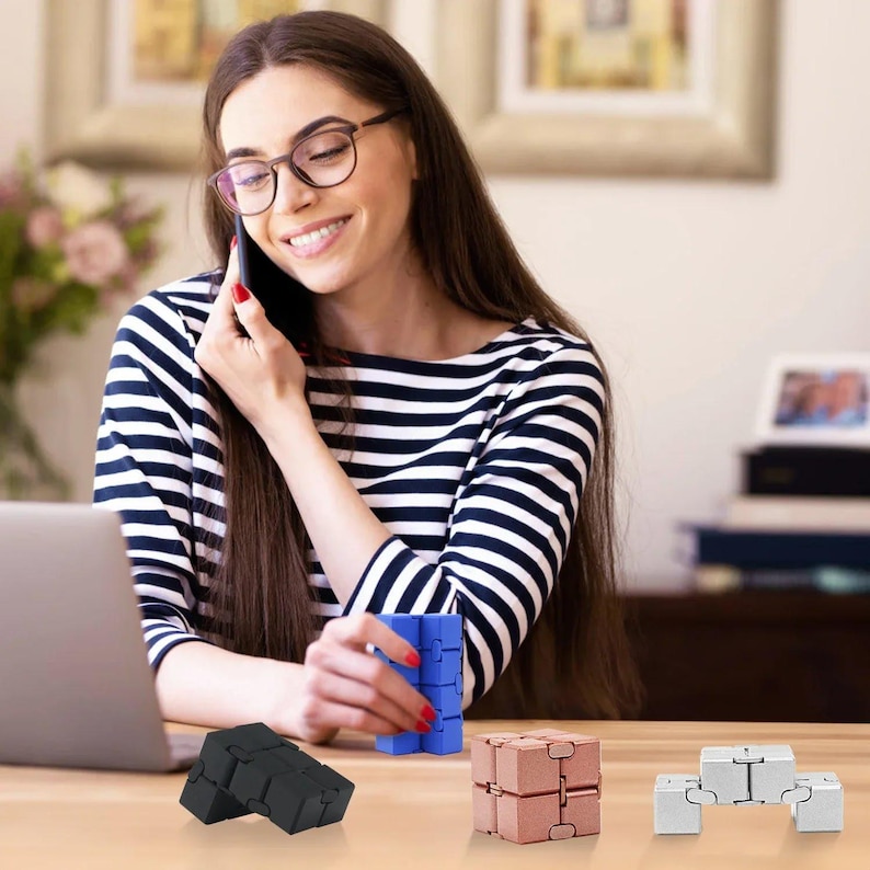 May include: A woman wearing glasses and a striped shirt is on a phone call. Several fidget toys in black, blue, rose gold, and silver are on the table. The toys are in various shapes and configurations.