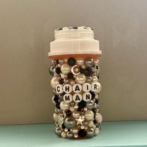 Puede incluir: Un frasco de pastillas decorativo cubierto con cuentas de varios tamaños en colores negro, blanco, plateado y perla. La palabra "CHAIRMAN" está escrita con cuentas de letras blancas. El frasco tiene una tapa blanca y está decorado con dijes de estrellas y planetas.