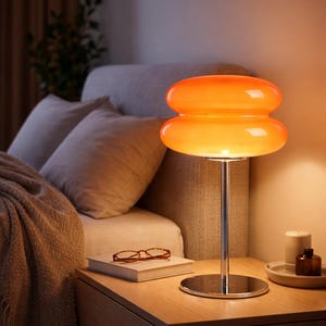 Peut inclure: Lampe de table en verre orange avec pied et base chromés. L'abat-jour est double et arrondi. La lampe est posée sur une table de chevet en bois, à côté d'un lit avec des oreillers et une couverture. Un livre et des lunettes sont également sur la table.