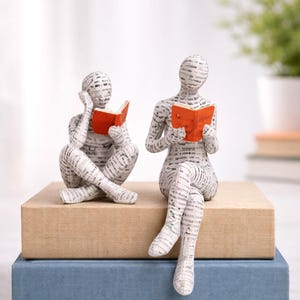 Peut inclure: Deux figurines lisant des livres avec des couvertures orange, faites d'un matériau semblable à du journal. Elles sont assises sur une pile de deux livres, un beige et un bleu, un objet décoratif sur le thème littéraire.