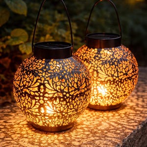 Peut inclure: Deux lanternes décoratives sphériques en métal noir avec des motifs floraux ajourés. Chaque lanterne est dotée d'une poignée et émet une lueur chaleureuse. Elles sont posées sur une surface en pierre, projetant des ombres.