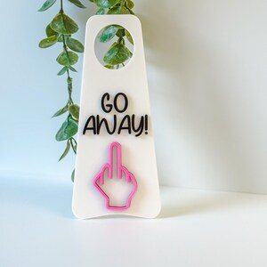 Könnte beinhalten: Ein weißer Türhänger mit dem schwarzen Text GO AWAY! und dem leuchtend rosa Umriss einer Hand, die den Mittelfinger zeigt. Der Hänger hat oben eine kreisförmige Aussparung zur Befestigung an einer Türklinke und eine glatte, flache Oberfläche.