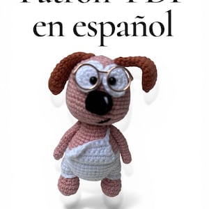 Pode incluir: Um cão de crochê usando óculos e macacão. O cão é principalmente rosa e branco, com orelhas marrons e um nariz preto. O texto "Patrón PDF en español" está no topo da imagem.