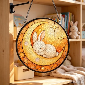 May include: A circular stained glass suncatcher featuring a white rabbit sleeping on a golden crescent moon. The design includes small star shapes and circular accents within a black frame, suspended by a black metal chain from a wall hook.