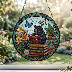 May include: A circular stained glass suncatcher featuring a black cat reading a book with a crescent moon on the cover. The cat sits on a stack of books labeled A Christmas Carol and Treasure Island, next to orange lilies and potted succulents.
