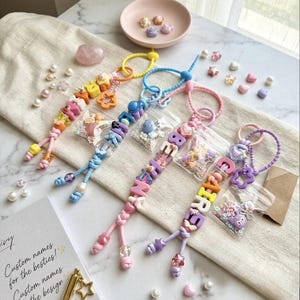 Könnte beinhalten: Bunte Schlüsselanhänger mit Namen, mit verschiedenen Anhängern und Perlen. Die Schlüsselanhänger sind in Rosa-, Blau-, Gelb- und Lilatönen gehalten. Kleine Beutel mit Perlen und Anhängern sind ebenfalls sichtbar, zusammen mit einer rosa Schale und verstreuten Perlen.