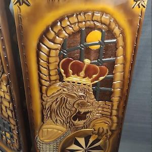 Könnte beinhalten: Eine rechteckige braune Wandplatte mit einem Relief eines Löwen, der eine Krone trägt. Der Löwe befindet sich vor einem steintexturierten Torbogen mit einem gelben kreisförmigen Mond, der durch ein dunkles Gitterfenster sichtbar ist. Unten ist ein Stern.