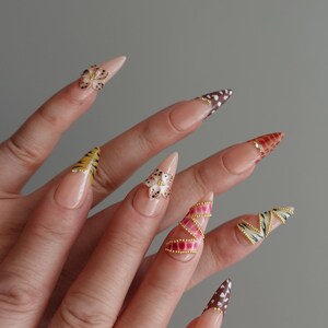 May include: A hand with long, pointed, artificial nails. The nails feature various designs, including floral accents, animal prints, and gold embellishments. The color palette includes nude, brown, yellow, pink, and white.
