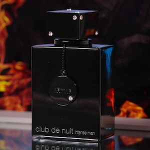 May include: A black rectangular perfume bottle with a square cap adorned with rhinestones. The bottle has the text "club de nuit intense man" printed on the front. A small, round charm with the brand name and a rhinestone hangs from the bottle.