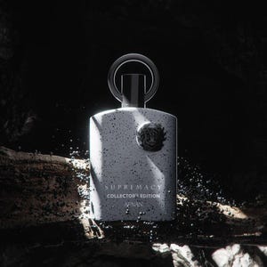 May include: A white perfume bottle with a speckled design, a black cap, and a silver ring handle. The bottle has the words "SUPREMACY COLLECTOR'S EDITION AFNAN" printed on it. The background is dark with a rocky texture.
