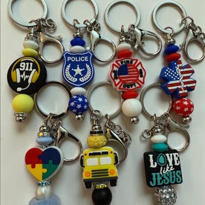 May include: A collection of keychains with various charms. Charms include a 911 logo, a police badge, a fire department emblem, an American flag, a puzzle heart, a yellow school bus, and a black rectangle with the text "LOVE like JESUS".