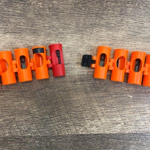 May include: A segmented toy ammunition belt featuring bright orange plastic cylindrical links with black internal details. One link is red and includes a black sliding switch mechanism. The belt is separated into two distinct sections.