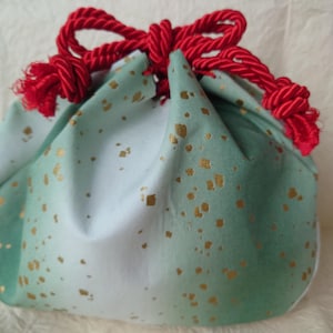 May include: A small, round fabric pouch with a drawstring closure. The pouch is a gradient of light blue to green, speckled with gold flecks. A thick, red rope forms the drawstring and bow at the top, adding a pop of color.