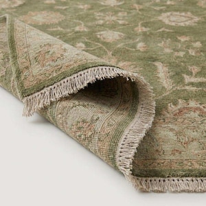 May include: A sage green area rug featuring a repeating floral pattern in muted beige and tan tones. The rug is folded over to reveal a textured surface and a decorative fringe edge along the border, showcasing the woven construction and pile.