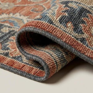 Puede incluir: Primer plano de una alfombra enrollada con un patrón detallado. La alfombra presenta una paleta de colores óxido, azul marino y beige, con un borde gris. El diseño intrincado sugiere un estilo tradicional o vintage, adecuado para la decoración del hogar.