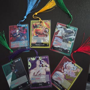 May include: Six rectangular trading card bookmarks encased in clear plastic sleeves, each featuring a One Piece character illustration, a power level of 5000, and a colourful silk tassel attached through a punched hole at the top centre.